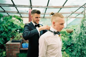 The groom helps one of his groomsmen put on his bowtie, hand-painted by the Groom's Mum taken by a film wedding photographer
