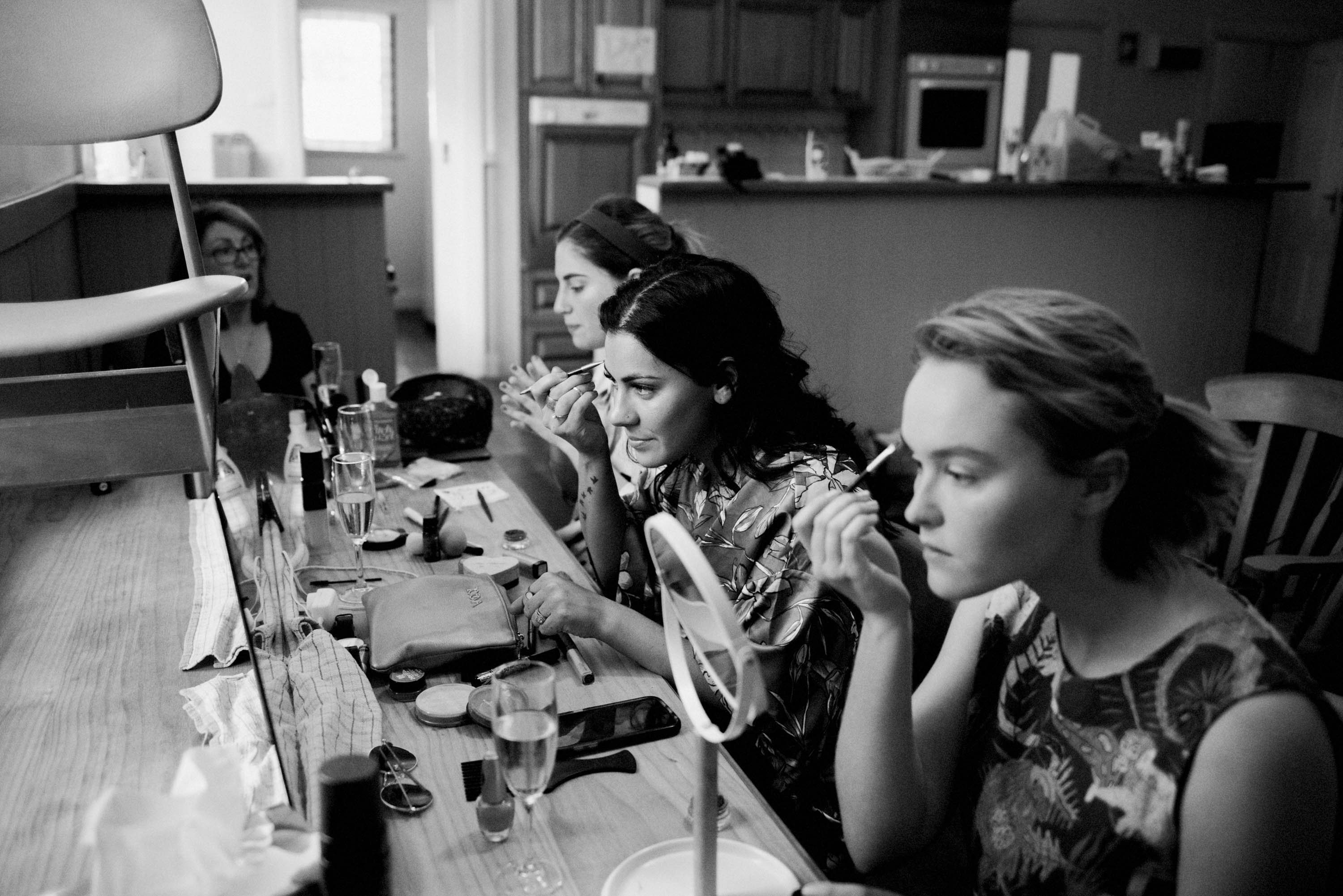 A black and white photo of bridesmaids doing their makeup at a long table.