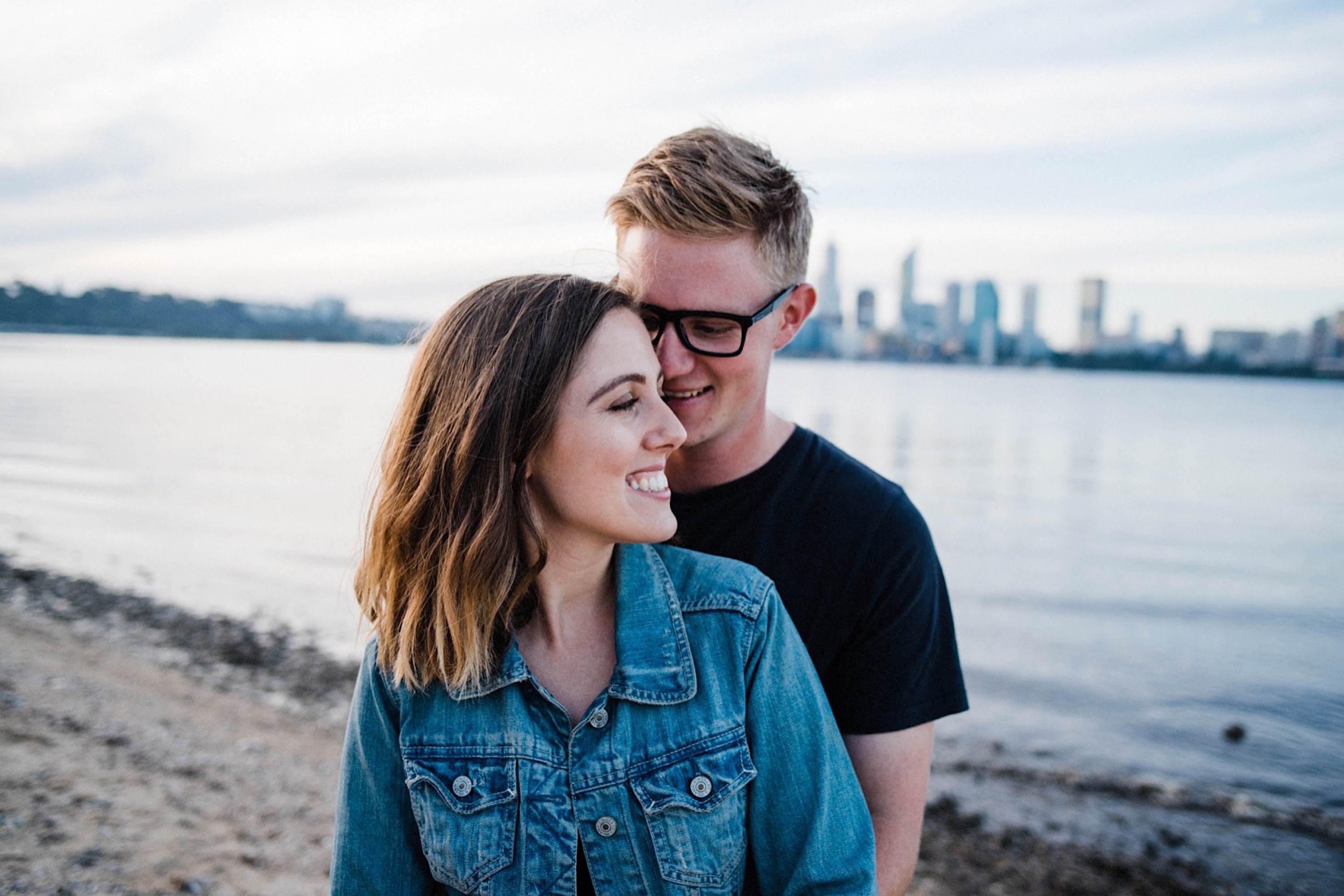 A photo of a young couple smiling together at Sir James Mitchell Park, looking over the Swan River and Perth City Skyline at sunset. 