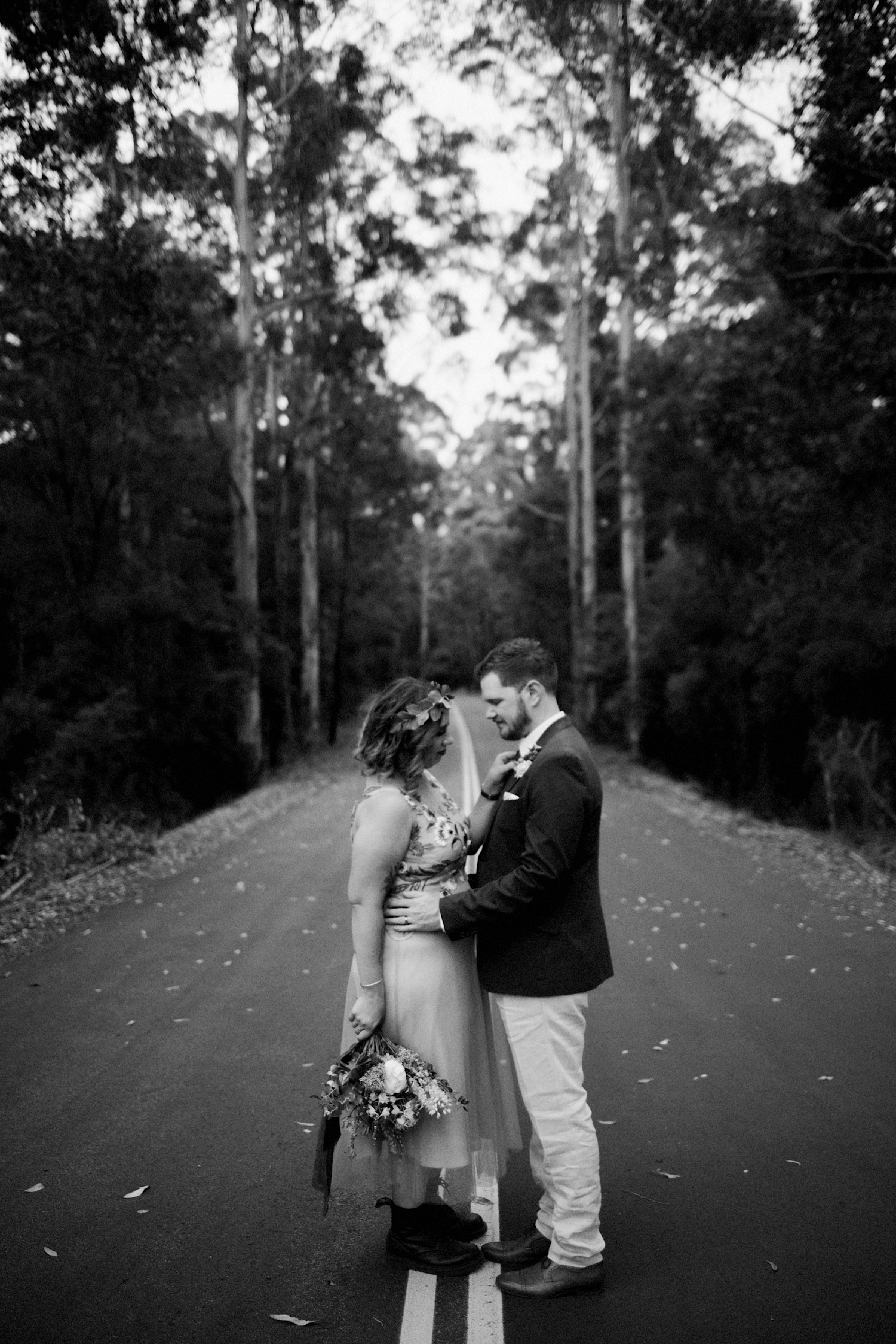 A black & white wedding portrait of the bride & groom on a country street surrounded by tall trees.