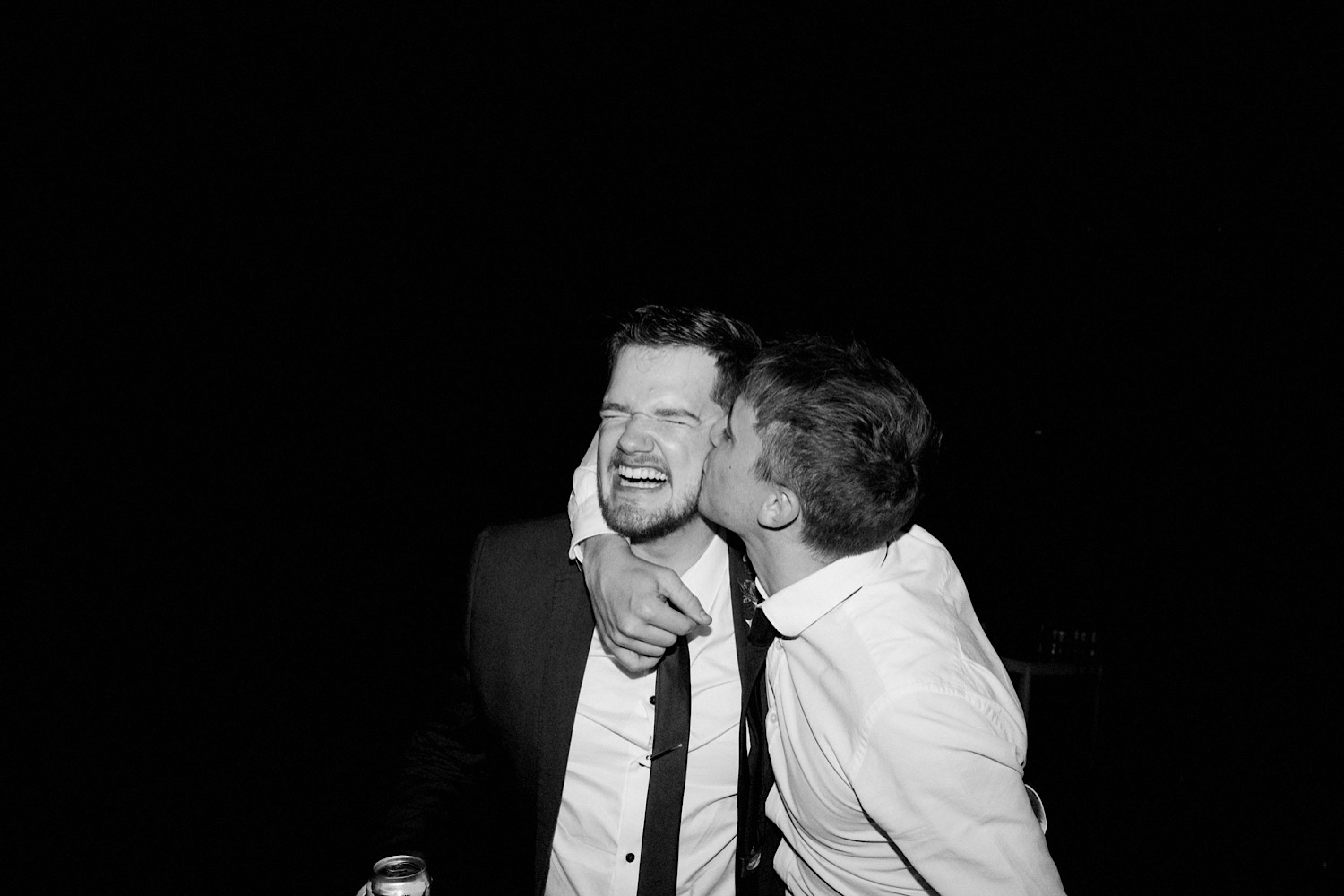 A black & white photo of the groom, who's full of joy, getting a kiss from one of his mates.