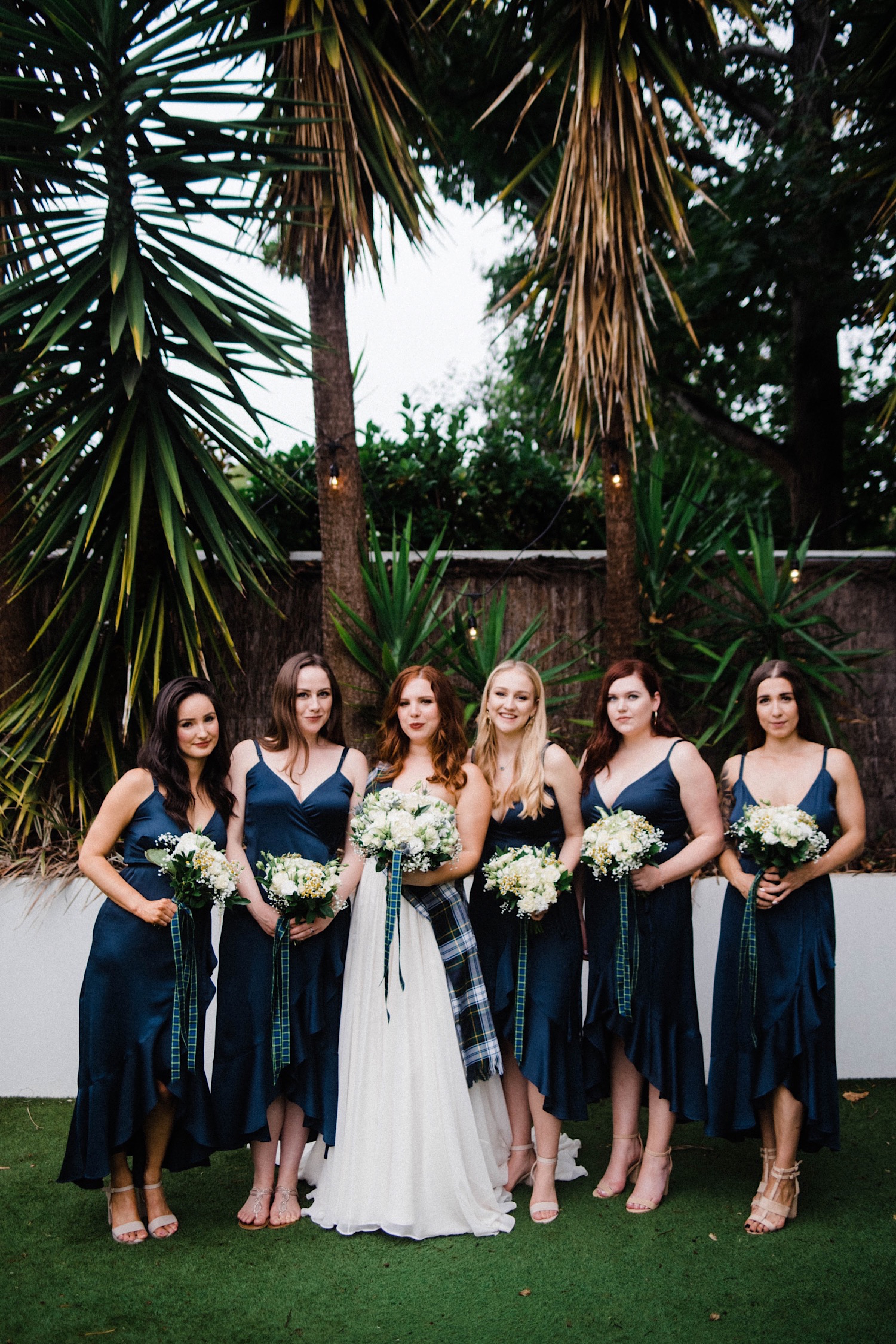 A portrait of the bride with her bridesmaid's at a Sustainable Backyard Wedding in front of a white wall and tall trees.