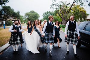 A documentary wedding portrait of the bride and her bridesmaids, the groom and his groomsmen who are wearing kilts, walking down a suburban street in the rain.