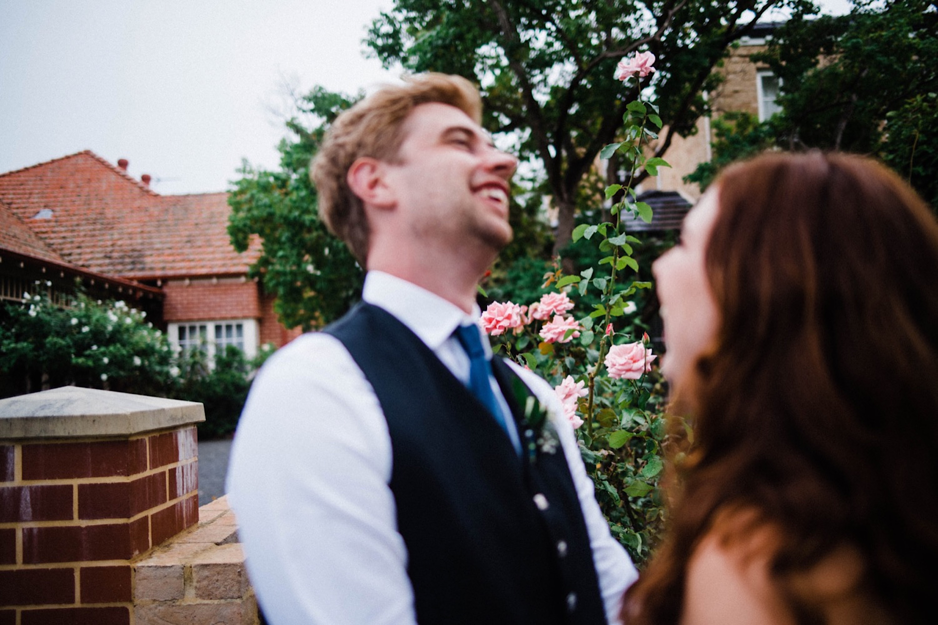 A candid wedding portriat of the groom leaning back and laughing in front of a rose garden.