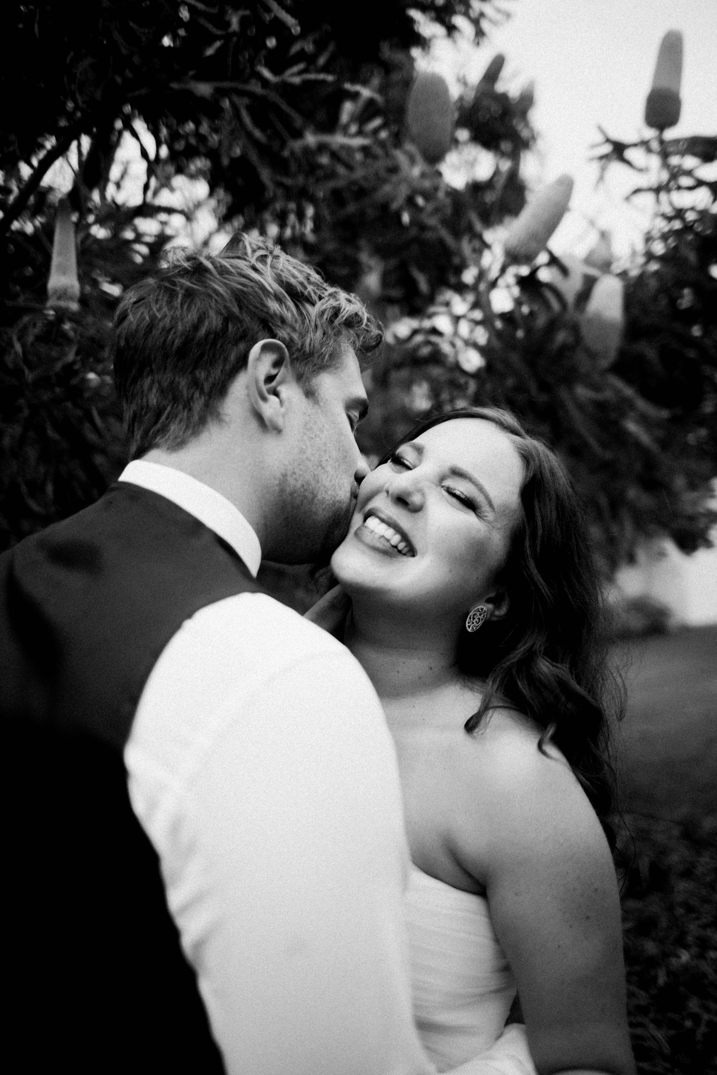 A black & white candid wedding portrait of the groom kissing the bride on the cheek while she laughs..