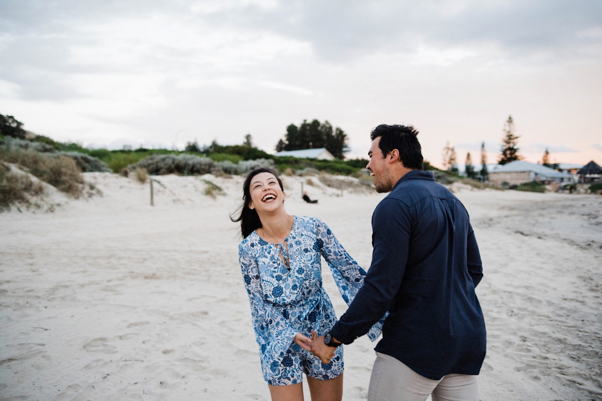 A Fremantle engagement photo taken at Bathers Beach of a couple laughing loudly and holding hands.