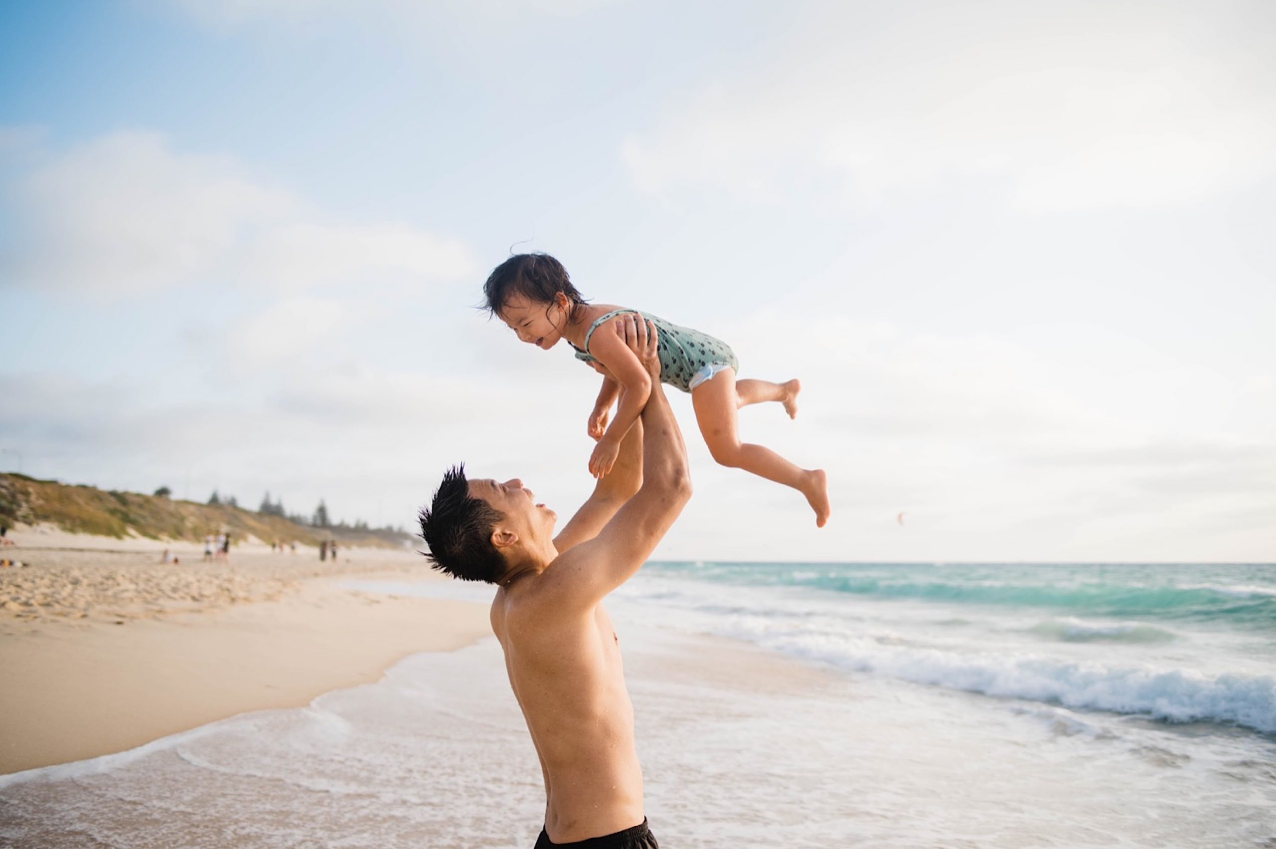 A photo of a Dad holding his toddler up above his head at the beach. They're both wearing bathers and laughing.