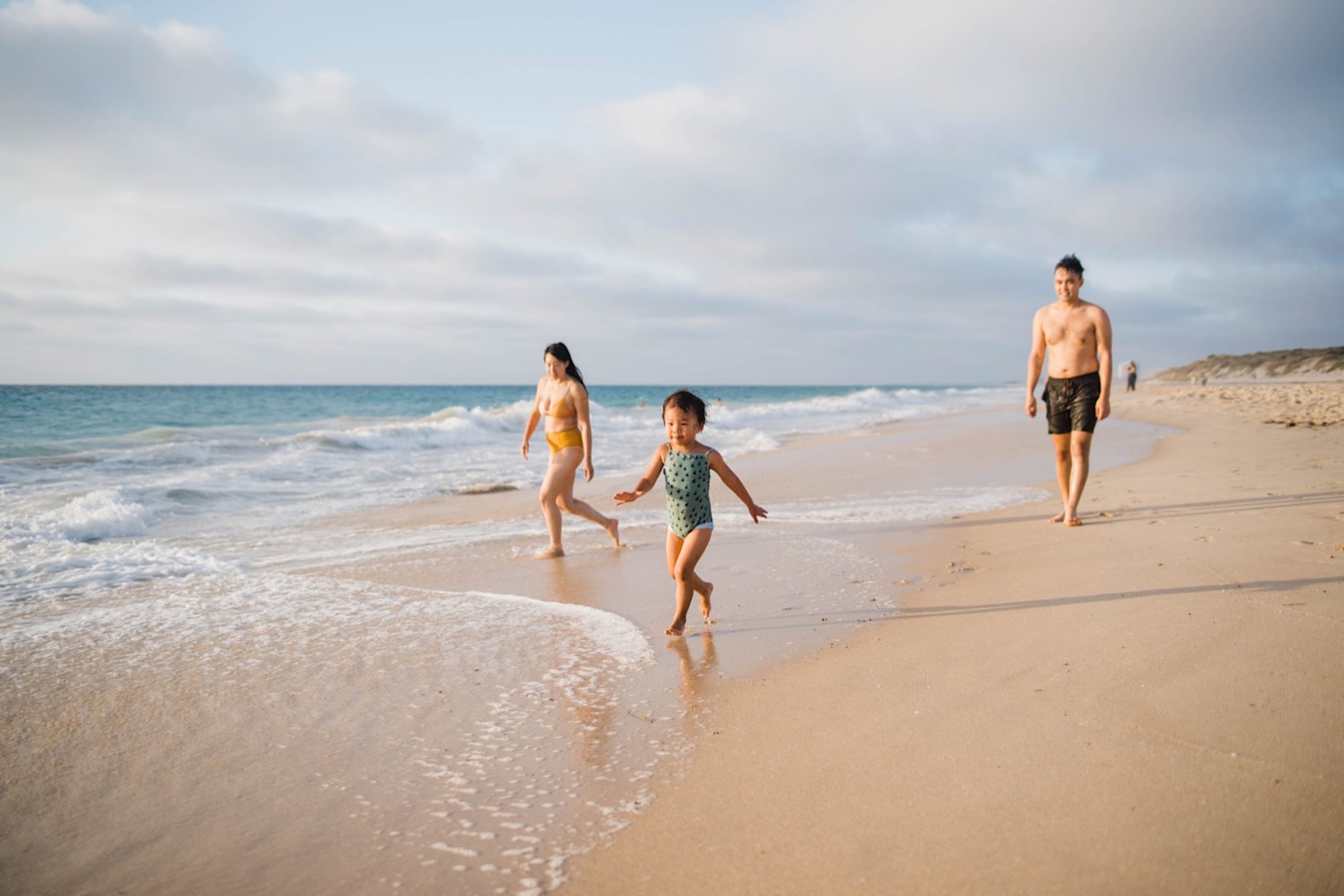 A photo of three people, one a toddler, walking along the beach and laughing as the little girl runs to the waves and then away from them again.
