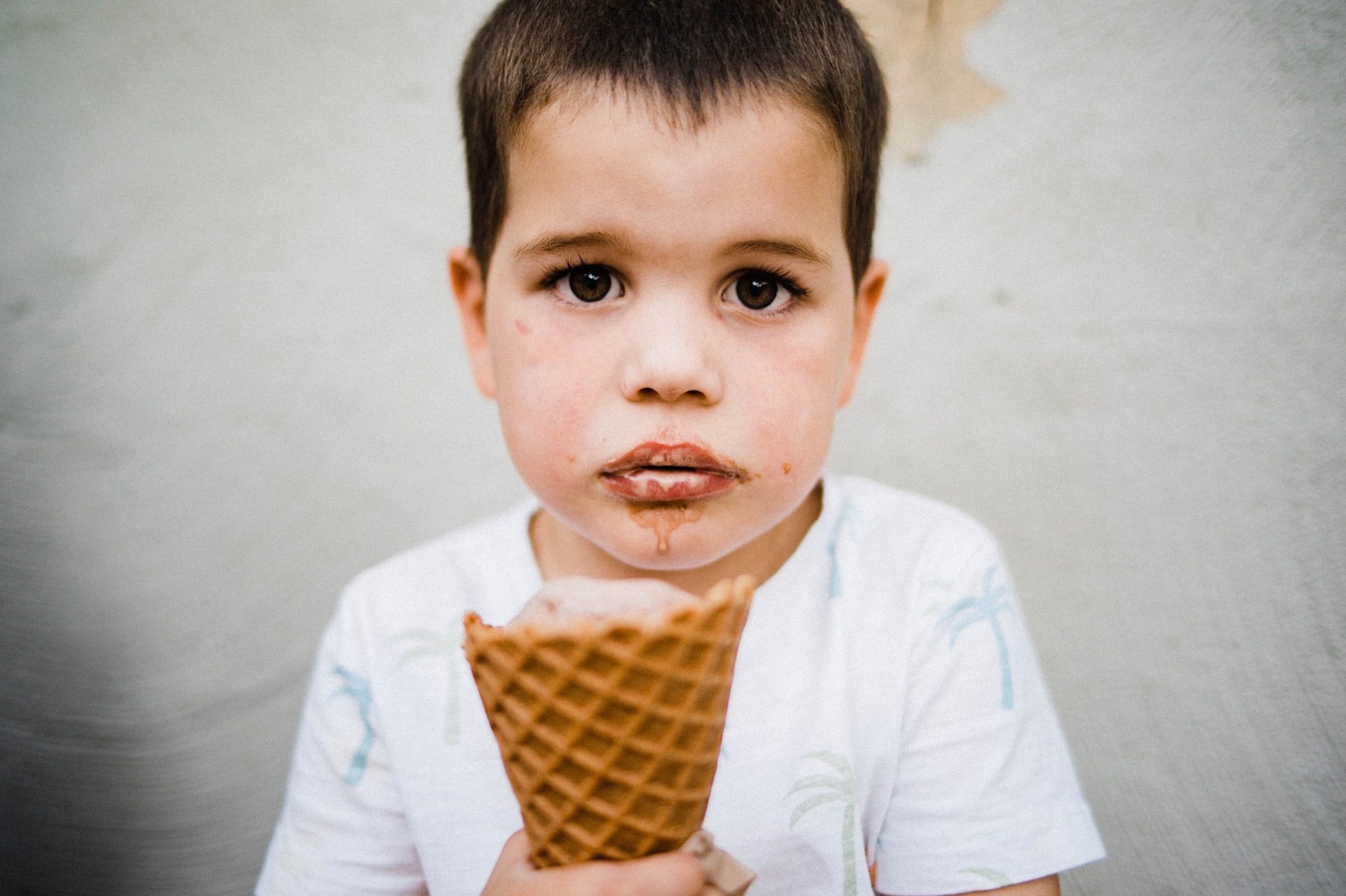 A close-up portrait of a young boy looking seriously at the camera, holding an ice cream cone, with ice cream on his lips and melting down his chin.