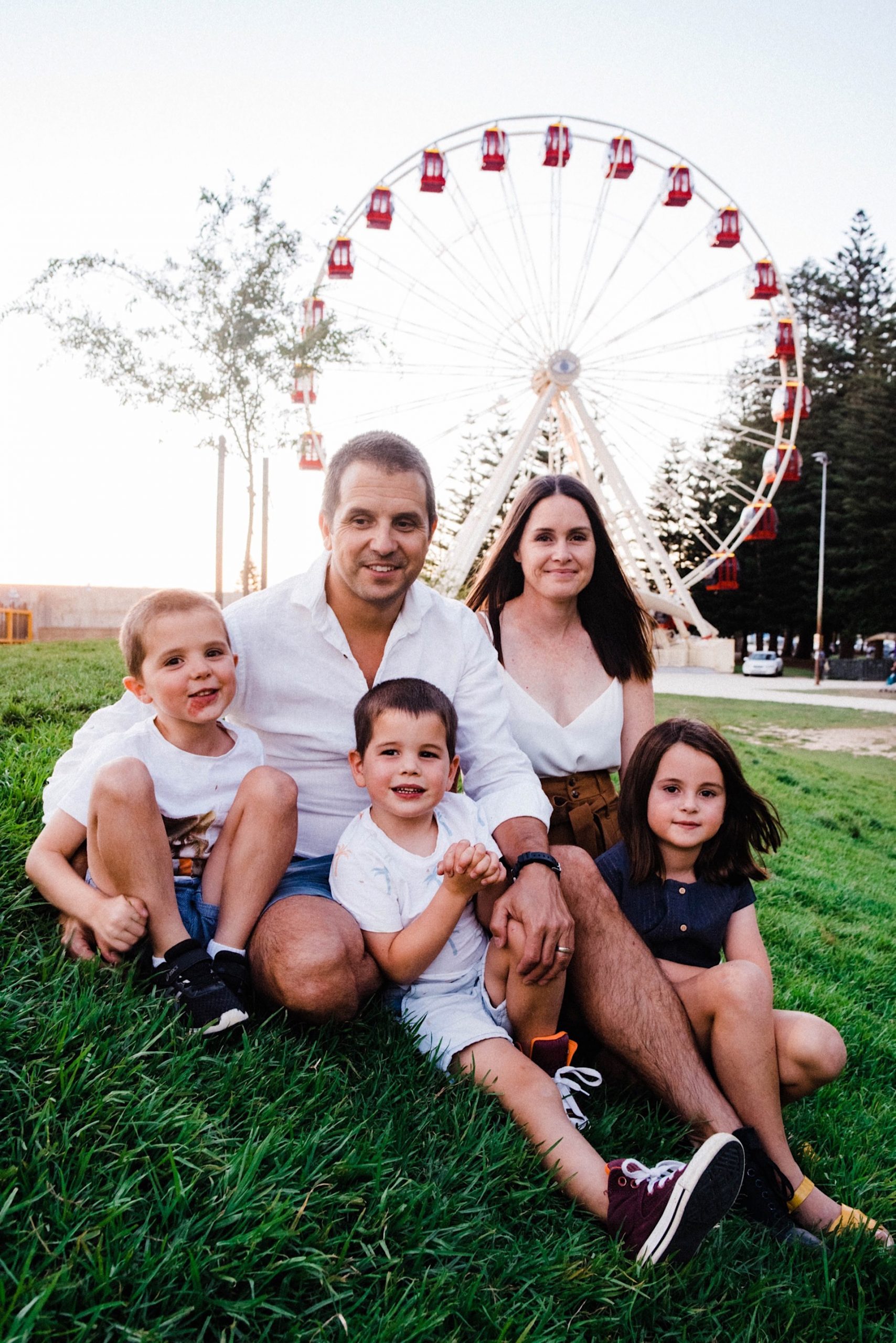 A portrait of a family of five sitting on a grassy hill at the Esplande Park, Fremantle, with a ferris wheel in the background.