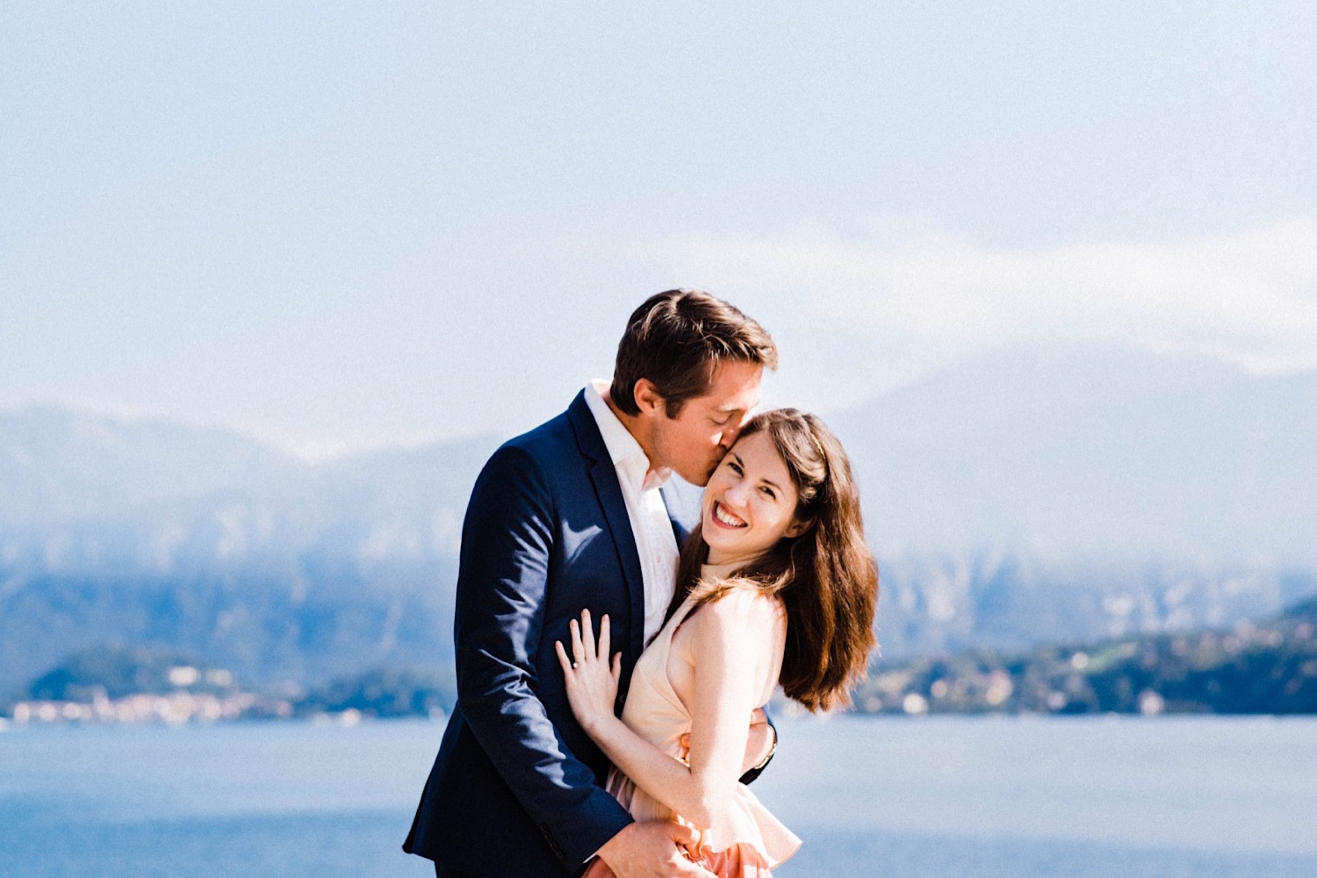 A couple stand in the middle of the frame, she smiles at the camera and he kisses her cheek. Lake Como and its surrounding mountains are in the background.