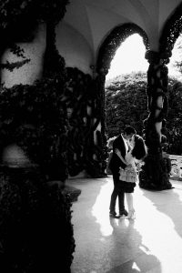 A black & white wide portrait photo of a couple standing in a patch of sunlight sharing a kiss.