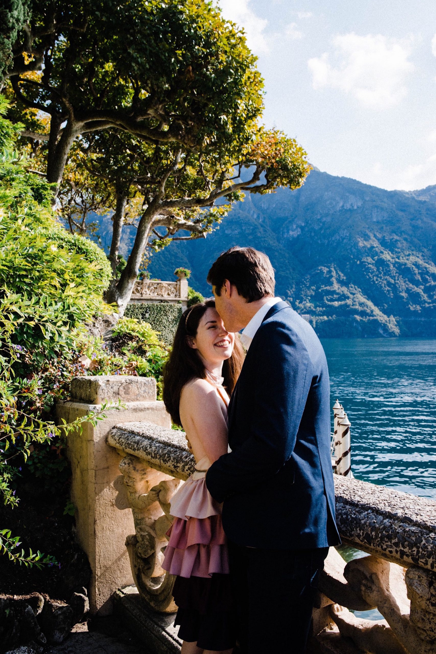 A portrait photo of a couple leaning against stone balustrades at Villa Balbianello, with gardens on one side and Lake Como on the other.