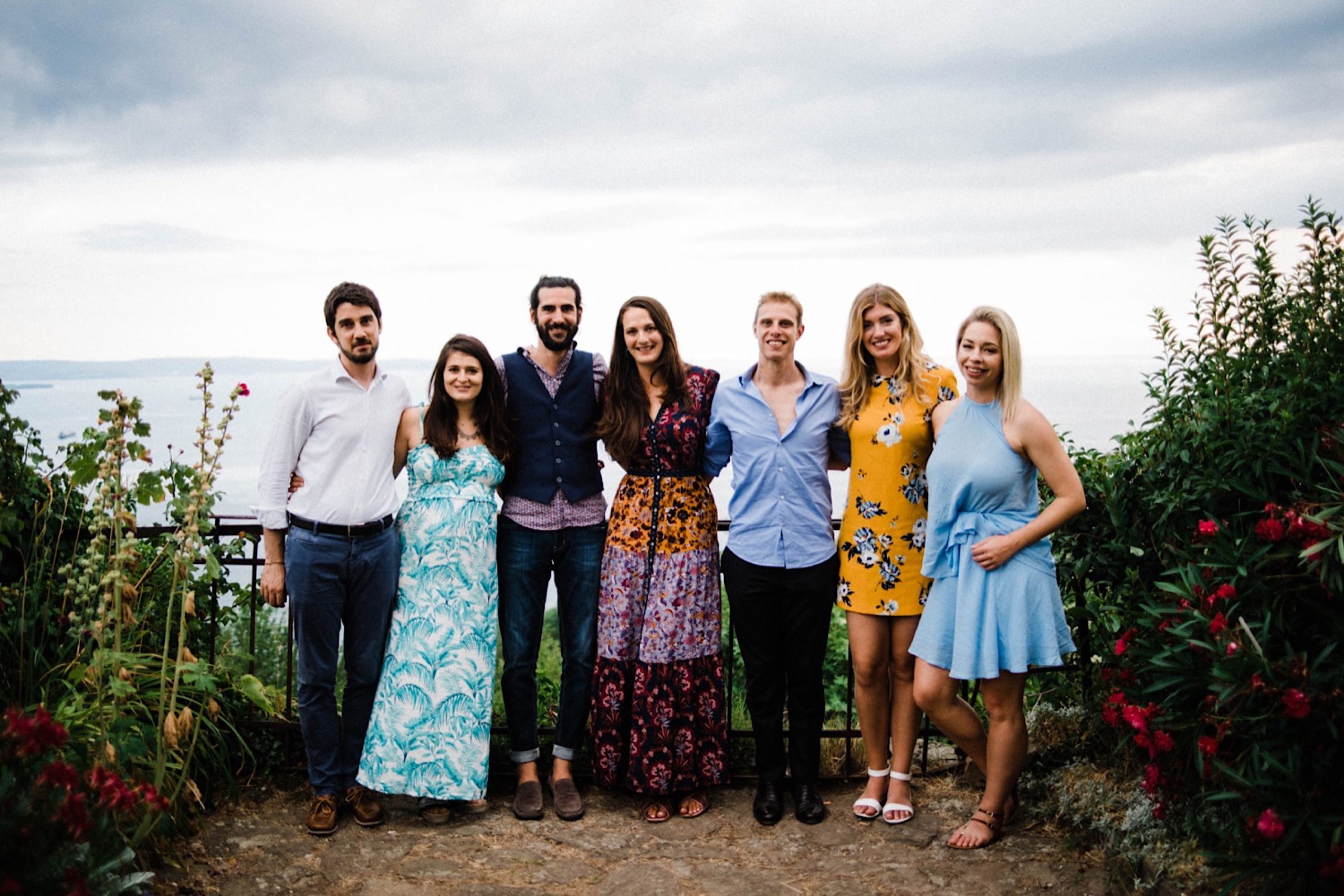 A group photo from an Intimate Destination Wedding in Trieste, Italy. The group wears colourful clothes, and stand in a garden looking out over the sea with stormy skies in the background.