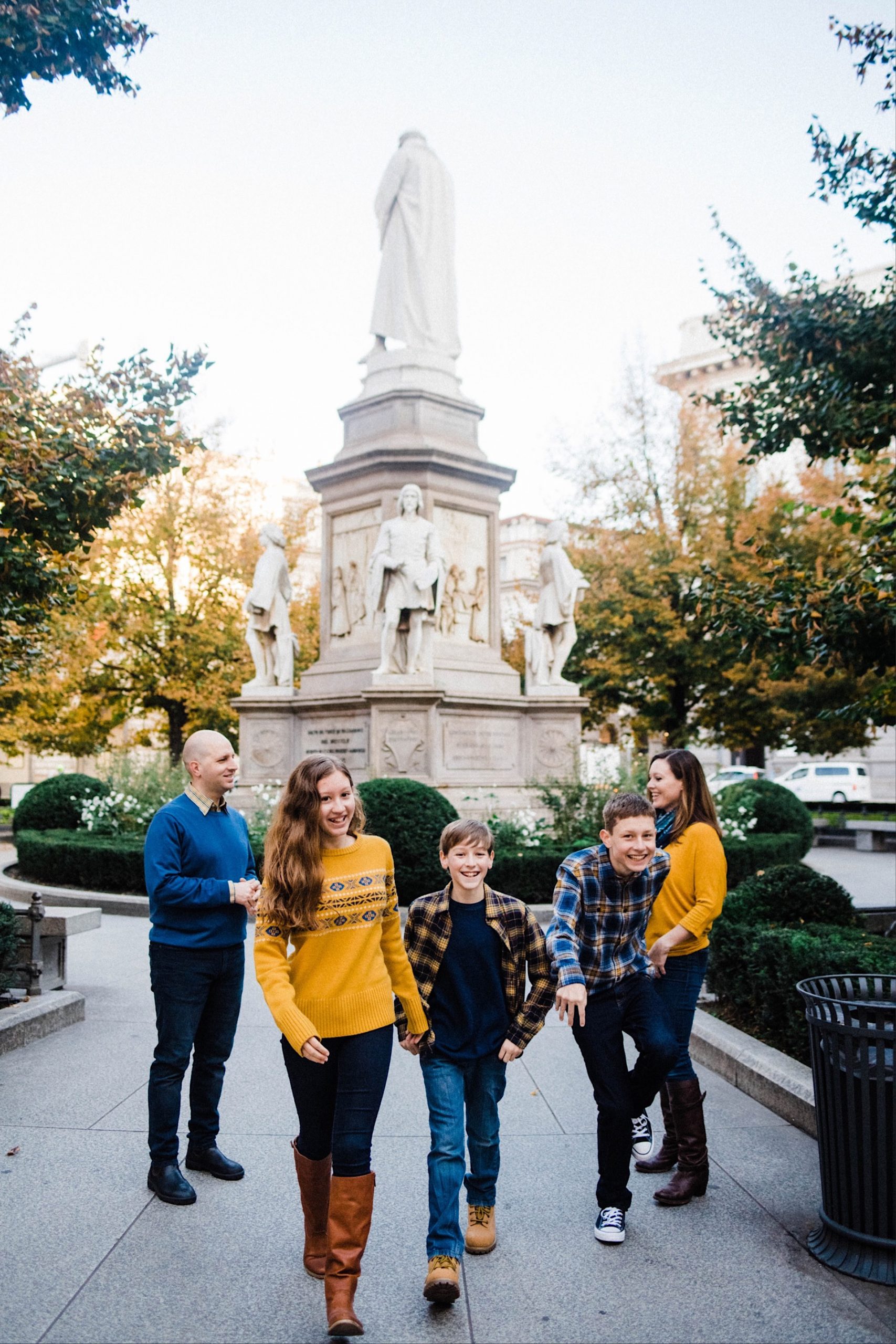 Wide portrait photo of three teenagers walking towards the camera while their parents laugh, taking at the Piazza della Scala in Milan.