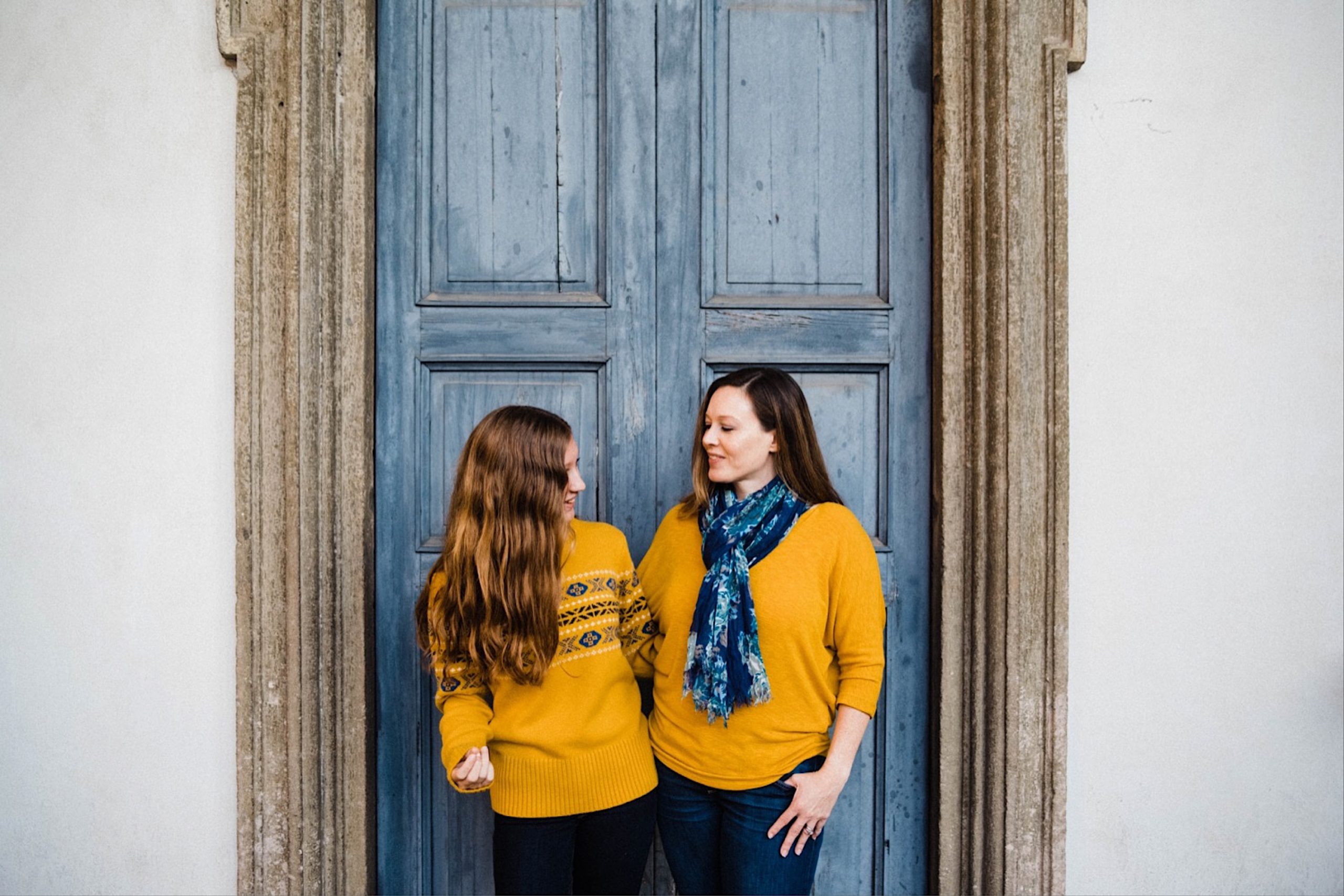 Portrait photo of a mother and daughter standing together, smiling. They both wear yellow sweaters and are standing in front of a blue door.