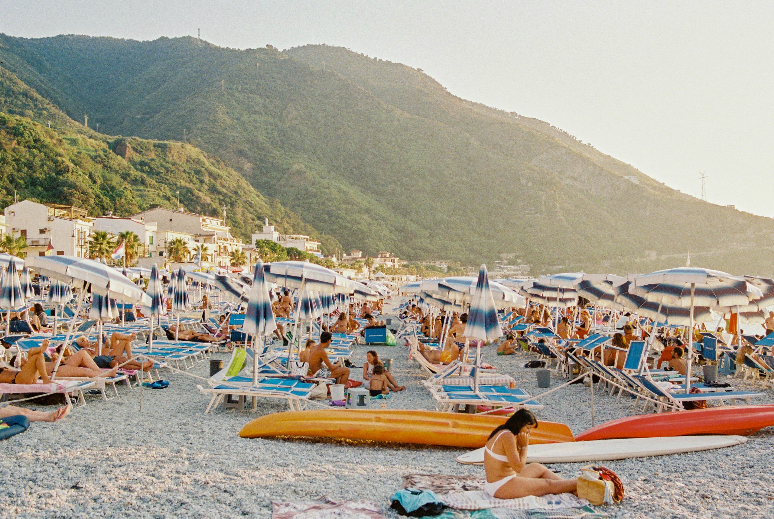 A 35mm film photo taken at Scilla Beach, in Calabria. In the foreground, a woman sits on her towel in white swimwear, behind her are endless rows of blue & white umbrellas, and behind that the a green hill covered in a golden haze from the sunset & salty water.