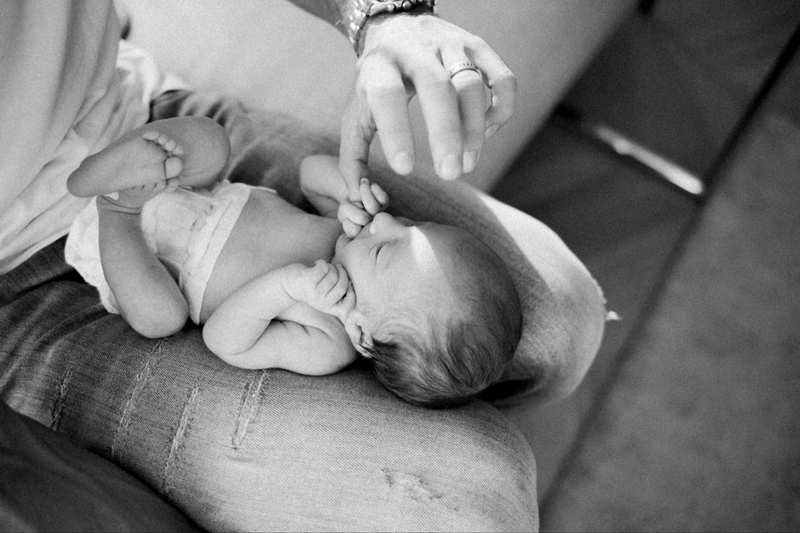 Black & white, Italy Newborn Photography of a newborn baby lying on her Dad's lap, holding his thumb in her hand.