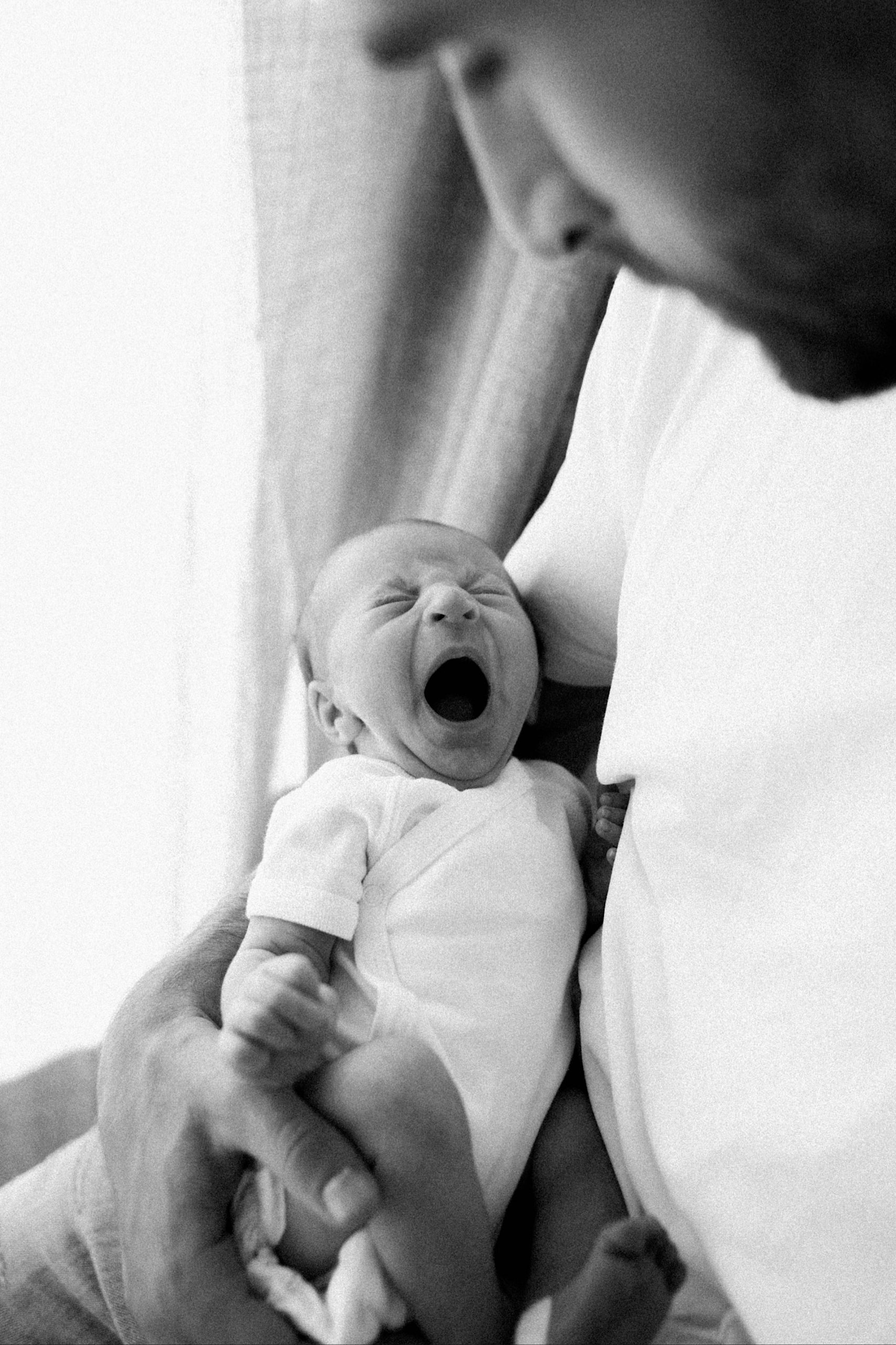 Black & White Italy Newborn Photography of a new baby yawning while in her Dad's arms, who is looking at her.