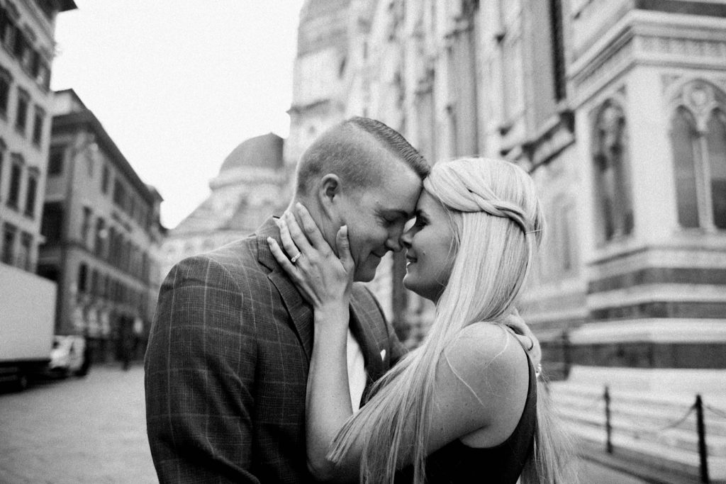 A black and white landscape photo of a couple standing forehead to forehead, with Florence's historic centre in the background.