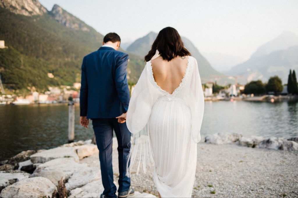 Romantic Boho Summer Elopement in Italy photo of a couple walking towards a lake with mountains in the background. The groom wears a blue May Faber Milano suit and the bride wears a backless, sustainable wedding gown by Leila Hafzi.