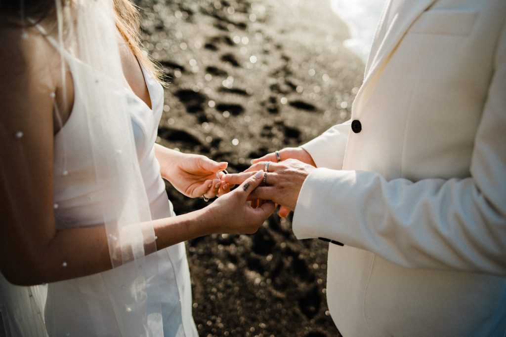 A close up shot of two people in wedding clothes exchanging rings, with black sand glinting behind them, to demonstrate How to Elope in Positano.