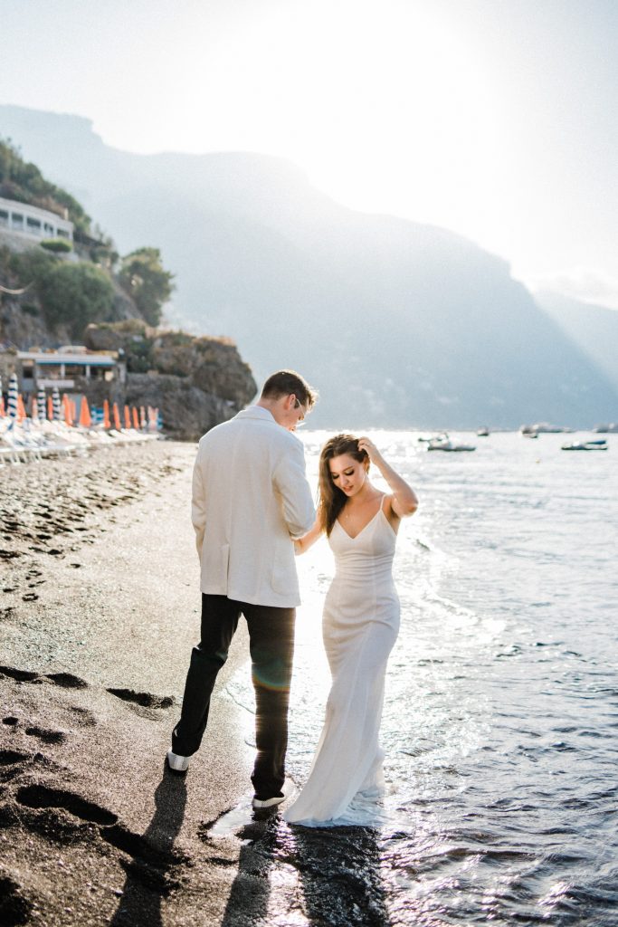 A portrait-oriented image, of a couple standing together in wedding clothes, her feet in the water, at the Spiaggia di Fornillo to demonstrate How to Elope in Positano.
