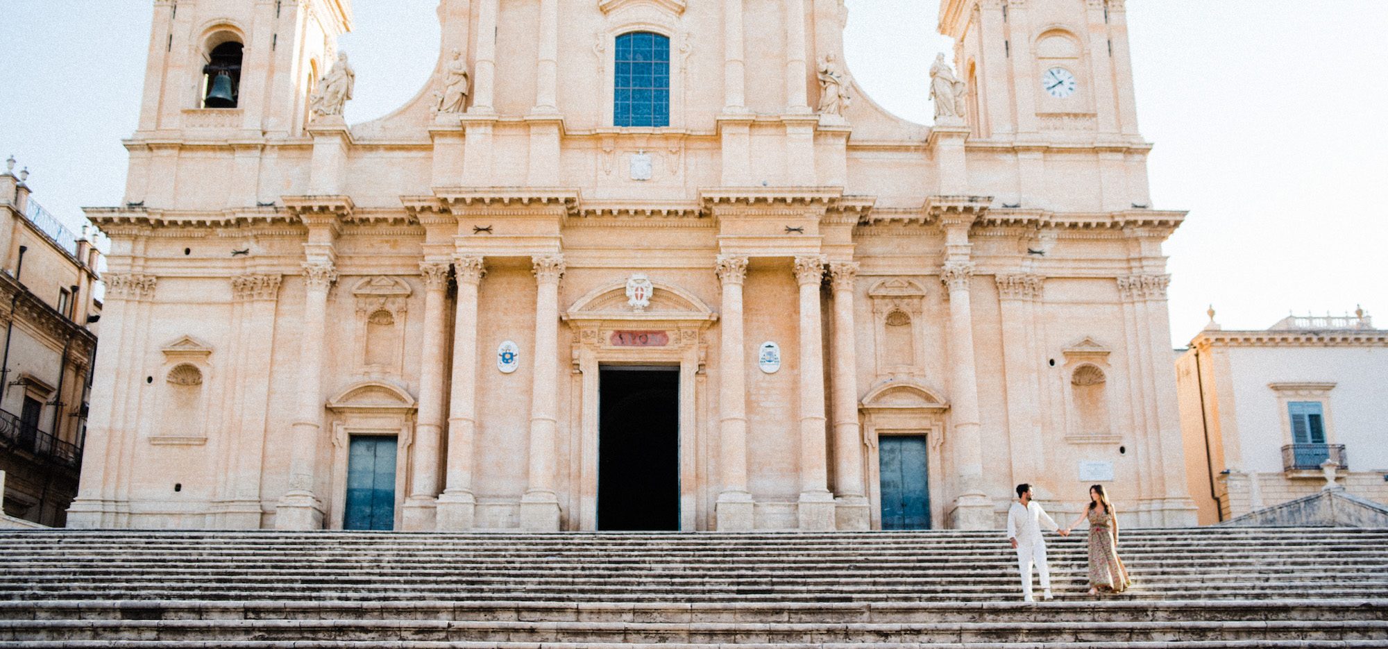 Wide shot of a couple holding hands and walking down the stairs in front of Noto Cathedral during their Noto Portrait Photography session.