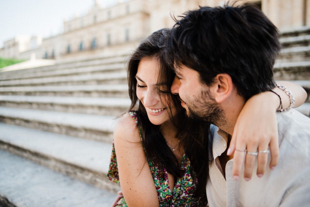 Close up image of a couple cuddling on the steps of Noto Cathedral during a Noto Portrait Photography session. She's smiling with her arm around his neck and he's resting his forehead on her hair.