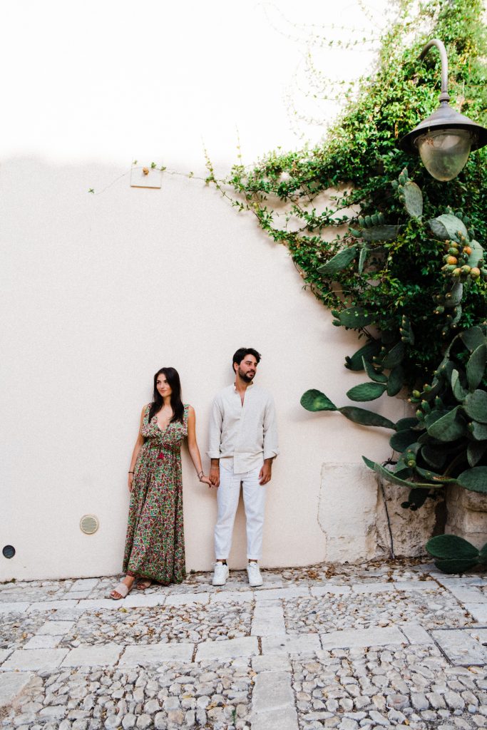Wide shot taken by a Photographer in Sicily of a couple standing hand-in-hand, leaning against a white wall with green vines and a prickly pear growing across the right side of the wall.