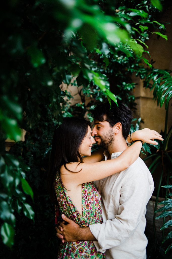 Medium shot of a couple resting their faces together, surrounded by greenery in a courtyard in Noto, Sicily. She's wearing a floral dress and he's wearing a white linen shirt for their Noto Portrait Photography session.