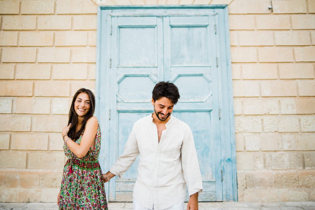 Candid image of a couple walking away from a limestone wall with an old powder-blue door in the centre. They're both laughing, he's looking down and she looks at the camera, during their Noto Couples Portraits.