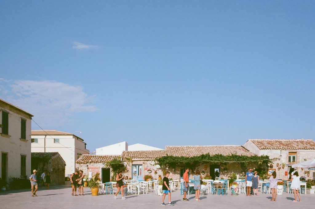 A landscape photo of Marzamemi, a great day trip location when you Visit Noto, Sicily. The top two-thirds of the photo are blue sky, the bottom shows a colourful piazza filled with blue & white chairs and people in summer clothes.