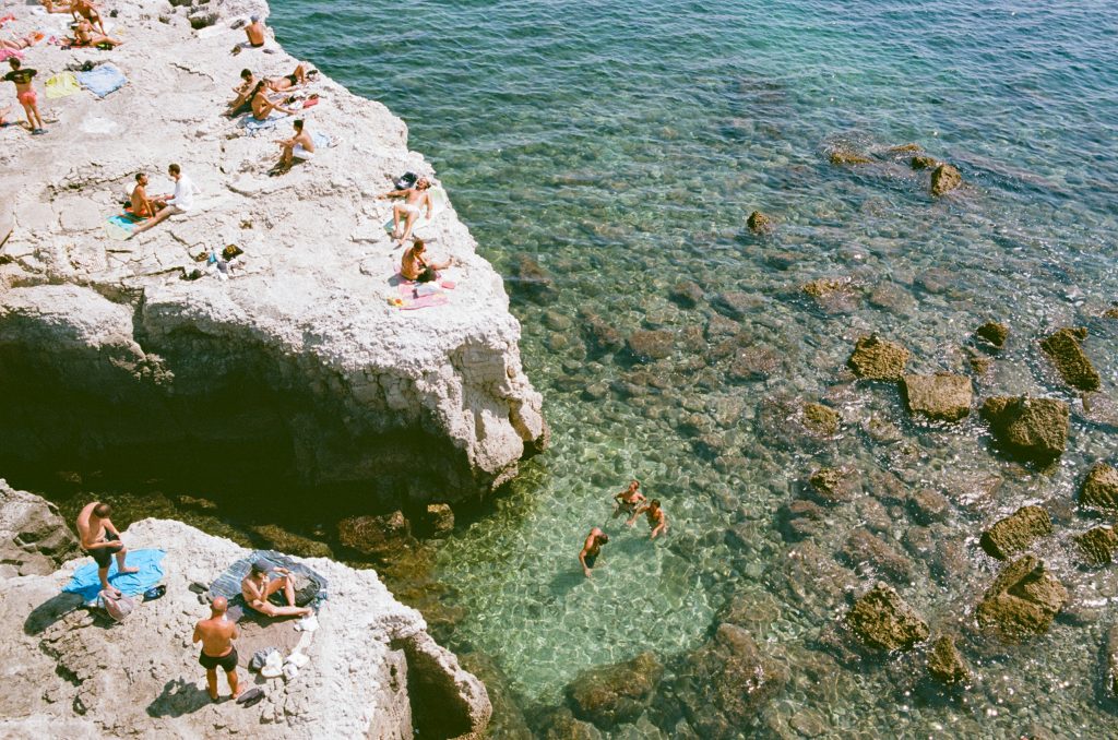 A photo taken from above of a popular swimming spot in Ortigia, Sicily. People lie on high white rocks to the left, while a few stand in the shallows of the sea to the right.